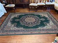 Full view of the rectangular green rug with floral border and medallion center in a room with wood floor and chairs.