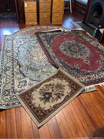 Three rugs displayed on hardwood floor showing size and pattern variations: a green-blue oriental rug, a red oriental rug, and a small floral decorative rug.