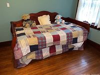 Full frontal view of wood frame daybed made in medium brown stain with patchwork quilt bedding, decorative pillows, and plush toys on bed.
