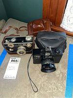 Three vintage cameras displayed on a table with their cases: Lavec LT-002, Argus with leather case, and Silette with black case.