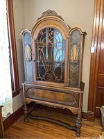 Front view of vintage wooden china cabinet with carved accents, glass door with scroll grille, and single drawer.