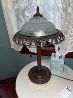 Full view of the vintage table lamp showing the metal base and the ornate metal shade with frosted glass insert against a lace curtain background.