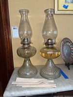 Pair of vintage glass oil lamps standing side by side on marble surface with extra replacement cotton wicks laid in front.