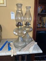Pair of vintage oil lamps with tall glass chimneys and fluted glass bases, showing included extra wicks and candle snuffer on marble surface.