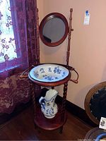 Full view of vintage wooden wash stand with round mirror, ceramic basin, and pitcher on wooden shelf.