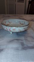 Side view of small round porcelain bowl with pink bridal rose pattern and gold trim on edge, showing three small feet