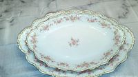 Two white oval serving platters with pink rose floral design and gold scalloped edges displayed flat on a marble surface.