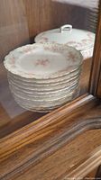 Stack of eleven small scalloped China plates with pink floral pattern and gold trim inside a wooden cabinet.