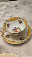 Tea cup and saucer set placed on a cloth surface showing top and inside of the cup with fruit and yellow decorative pattern.