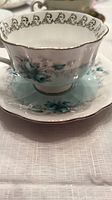 Close-up of the tea cup and saucer showing turquoise rose floral pattern with gold rim and ornate cup handle.