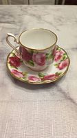 White tea cup and saucer set with pink rose floral design and gold trim, displayed on a white textured tablecloth.