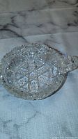 Clear crystal display bowl with intricate cut-glass patterns and a handle, shown on a light cloth surface.