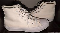 Side view of white high-top Converse Chuck Taylor All Star II shoes showing pair of shoes side by side on dark surface. White canvas with matching white laces and toe caps shown.