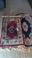 Two area rugs shown together on a larger beige carpet: one rectangular multicolored red and beige floral motif rug with fringes, one oval dark green and beige floral medallion rug with fringes.