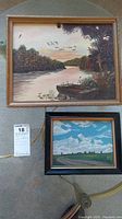 Two oil paintings on board shown in a photo on a carpeted floor. One painting shows a river with a boat and trees at sunset, the other a rural landscape with road and clouds.