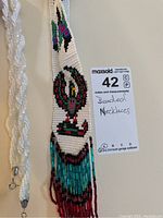 Clear photo showing close-up and details of two necklaces with different bead styles, one indigenous design with multi-colored beads and fringe, one twisted white beaded necklace.