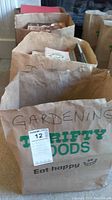 Photo of four brown paper bags filled with books. The front bag is labeled 'GARDENING' and is from Thrifty Foods store. The bags are full of assorted books visible from the top.