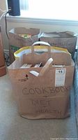 Four brown paper bags filled with assorted books. One bag labeled 'Cookbooks + Diet + Health' in front, and two bags labeled 'Mystery/Thrillers' in the back near a window.