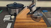Photo of stainless steel Crockpot with lid, wooden knife block holding two knives, glass saucepan with lid and black handle, white handheld milkshake maker, and small white timer device all arranged on a countertop near a wooden cutting board.