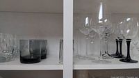 Shelves showing various sorts of clear wine glasses, some with black stems, alongside clear tumblers and decorative glass containers.