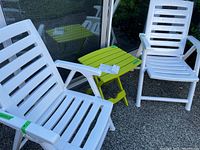 Two white resin folding chairs with slatted backs and seats, positioned on either side of a bright green side folding table with slatted top outdoors on concrete.