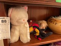 Four plush toys displayed on a wooden shelf including a white cat, a toucan, and others, alongside a yellow ceramic bowl and sea shells.