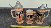 Three vintage beer steins displayed on a counter. Two have pewter lids and one is lidless. All have colorful relief artwork and ornate handles.