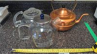 Three teapots: copper teapot, clear glass teapot with blue knob lid, and vintage Pyrex glass teapot, all placed on countertop with measuring tape.