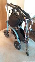 Wheeled walker showing padded seat, large wheels, and attached storage bag with two colorful canes hanging on it.