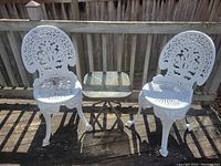 Full view of two white cast metal bistro chairs flanking an outdoor side table with tempered glass top on a wooden deck.
