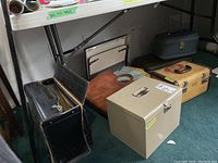 Image showing multiple briefcases and travel cases on floor under a table, including black suitcase, brown leather file case, metal file case, and blue toiletries travel case.