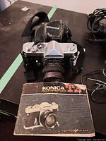 Konica T3 camera with lens and worn black Konica case resting on table, including Konica T3 user manual booklet.