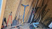 Overall view of assortment of loppers, saw, shovels, cultivator, rake, and edging rolls against plywood wall