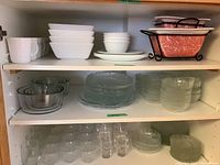 Shelves containing white Corelle dinnerware including mugs, square bowls, dinner plates, and glass dinnerware including various sized bowls and plates arranged neatly.