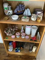 Photo of three wooden shelves with various decorative items including milk glass vases, model ships, seashell-covered boxes, candle holders, and candles.