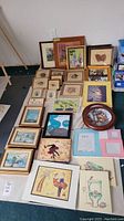 Overview of the collection showing multiple vintage framed prints, needlepoint art, a wooden framed owl mirror, collector plates, and various small framed artworks placed on a table.