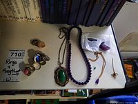 Overview of rings, amethyst bead necklace, green stone pendant necklace, and small amethyst pendant necklace on a table.