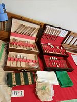 Two wooden cases containing 1881 Rogers silverware flatware sets displayed laid open showing knives, forks, spoons, and some serving pieces. Fabric napkins in green, white, and patterned styles are placed in front.