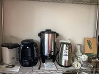 View of the lot showing the corn popper, Tassimo coffee maker, coffee urn, electric kettle, and small appliance parts on a metal shelf.