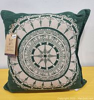 Square green cushion with intricate circular beige and dark green design and a hanging branded tag","reason":"To show cushion pattern and condition