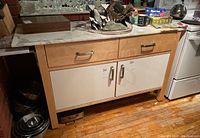 Full view of kitchen cabinet showing marble top, light wood frame, two drawers and two white cabinet doors with metal handles, placed on wooden floor next to stove and brick wall.