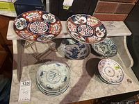 Wide shot showing two Imari plates on stands, blue and white scenic plate on stand, stacks of small bowls and small plates.