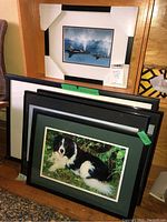 Stack of framed prints and photographs against wall, showing multiple items in lot including dog photo and whale print