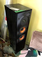 Photo showing one black tower speaker with orange cone midrange and woofer drivers, placed near a wall and a chair, with visible wear and scratches.