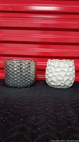 Front view of metal finish hexagon design pot and off-white beige textured pot side by side on black surface with red background.