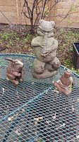Three garden figurines arranged on a metal table: a tall concrete frog playing a pipe, a small rabbit missing one ear, and a seated Buddha figurine.
