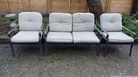 Three piece patio set with loveseat and two chairs arranged outside on stone pavement. Metal frames in dark brown finish with taupe cushions showing minor wear.