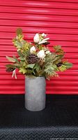 Artificial floral arrangement in a fluted galvanized metal pot against a red background. The foliage includes green leaves, white buds, brown pinecones, and lotus pods.