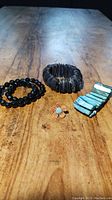 Top view of all five costume jewelry pieces displayed on wooden surface: two black plastic bead bracelets, one Mexican shell bracelet, one turquoise rectangular piece bracelet, and one flower ring.