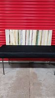 Full view of the abstract textured wall art panel against a red background and resting on a black bench, showing the vertical multicoloured stripes and texture.